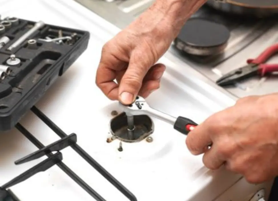 Technician using a wrench and screwdriver to repair a gas stove burner with a tool kit nearby.